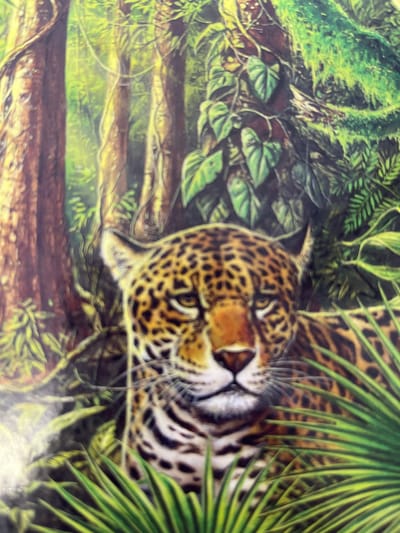The image is a close-up of a jaguar's face, rendered in a realistic painting style. The jaguar has characteristic spotted fur in shades of yellow, black, and white, and its eyes are focused forward. The background depicts a lush, green jungle environment with dense foliage, vines, and large tree trunks. The lighting suggests a dappled sunlight effect, common in forest canopies. There is no indication of any urban environment, including the city of Zhigalovo, Russia. The image solely focuses on the wildlife and its natural habitat.