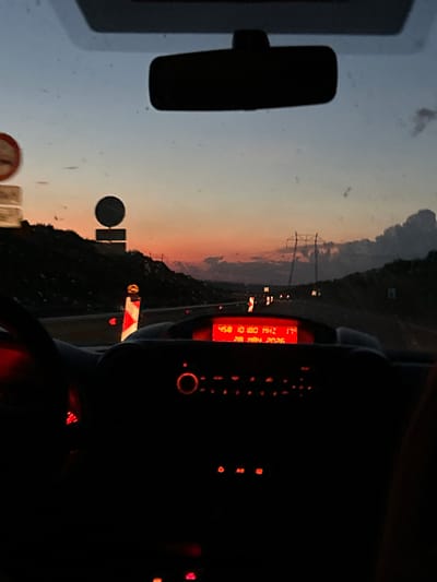 A view from inside a vehicle, looking out at a highway near Vinarsko, Bulgaria at dusk. Power lines stretch along the roadside with a sunset sky. Road signs indicate construction or maintenance work. The dashboard of the vehicle is visible, with a red display showing numbers and letters. The rearview mirror is also visible. No people are apparent.