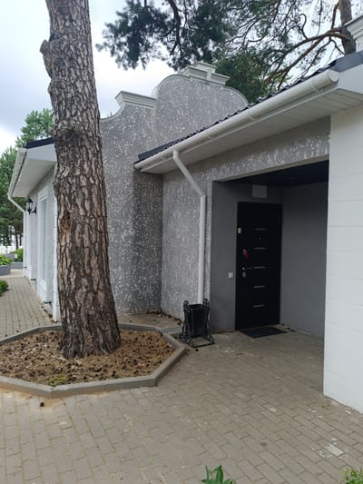 The image depicts the exterior of a building in the Старобінскі сельскі Савет, Belarus. A large tree trunk with rough bark is positioned prominently in the foreground, to the left of the frame. The tree is surrounded by a hexagonal concrete planter filled with pine needles and pine cones. The building features a textured gray facade with decorative white trim and a black roof. A white gutter system runs along the edge of the roof. To the right, a dark gray recessed entryway leads to a black door with horizontal metallic accents. A red key fob is attached to the door handle. A black doormat is placed in front of the door. A black metal waste bin with an ornate design stands near the entryway. The ground in front of the building is paved with light gray interlocking concrete bricks. In the background, to the left, glimpses of a white building with windows and a black outdoor lantern are visible. The environment appears to be a residential or commercial area with a predominantly overcast sky.