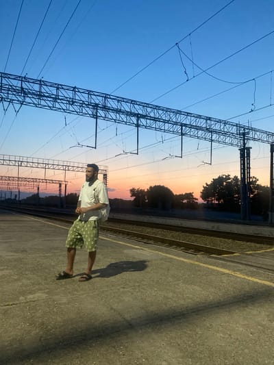 A man stands on a concrete train platform at dusk in Samur, Russia. He is wearing a white t-shirt, green patterned shorts, and dark sandals, with a white backpack on his shoulders. The sky is a gradient of deep blue to orange, indicating the sunset. Overhead, a network of power lines and metal gantries stretches across the frame. Train tracks run parallel to the platform, disappearing into the distance. Silhouetted trees and a low building are visible beyond the tracks. The lighting suggests natural light from the setting sun.
