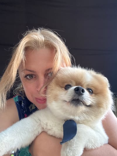 A woman with blonde hair and a dark floral top is holding a Pomeranian dog. The woman is looking at the camera with a slight smile, and her hair is partially covering her face. The dog is fluffy and light brown and white, with a blue bow around its neck. The background is dark, suggesting an indoor setting or evening time. The image does not contain any information regarding its location in Alakhadzi, Abkhazia.