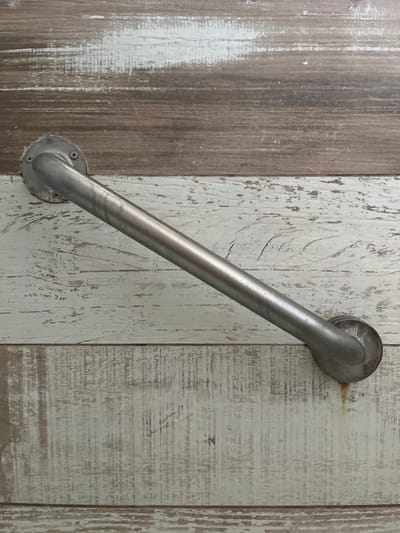 A silver grab bar is mounted on a wall featuring a wood panel design. The paneling alternates between light and dark faux wood grains with visible distress marks. The grab bar is angled, with circular bases at each end secured to the wall with screws. The overall aesthetic suggests a bathroom or mobility-assisted space with a rustic or farmhouse-inspired design.
