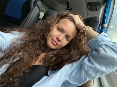 A woman with long, curly brown hair and light blue eyes is taking a selfie in what appears to be a vehicle. She is wearing a light blue shirt and a black top. Her right hand is resting on her head. A gold bracelet with pearls is visible on her right wrist. The background shows a black car seat with blue accents, a seatbelt, and window curtains, suggesting the interior of a bus or van. The lighting indicates it is daytime. The location is not specified in the image.