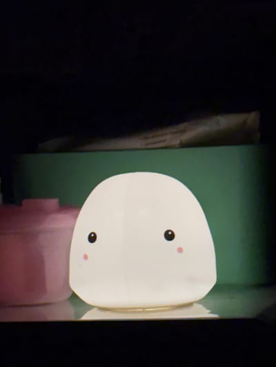 A white, egg-shaped night light with a cartoon face, consisting of two black dots for eyes and two small pink circles for cheeks, sits on a reflective surface. Behind it, a dark green rectangular object is visible, and behind that, a light-colored object with text, possibly a package, is sloped. To the left of the night light, a pink cylindrical object is partially visible. The background is dark, suggesting a dimly lit room or nighttime setting. The location is Sovkhoza "Razdol'e", Russia.