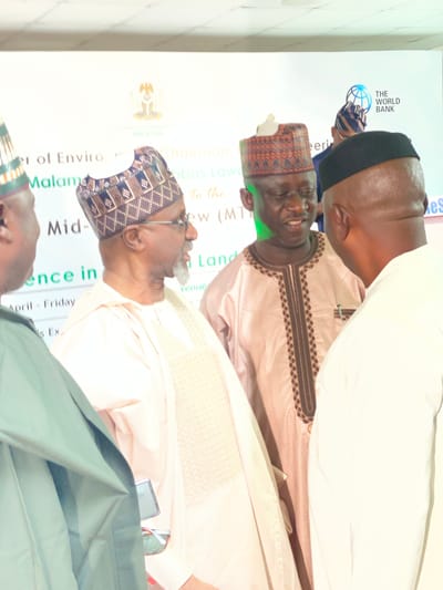 The image captures a scene from an event held in Kwarau, Nigeria. Three men are prominently featured in the frame. Two of the men are facing towards each other. The man on the left is dressed in a traditional attire with a patterned cap, while the man on the right is wearing a black cap and a patterned garment. Behind them, part of a banner is visible, mentioning “er of Enviro”, "Malaman", "Mid-", and "ence in". The banner also displays the logo of the World Bank and the coat of arms of Nigeria. Other text fragments, such as "To the," "Land," and "Venue" can be seen on the banner, but are obscured. The date "April - Friday" is also written, suggesting the timing of the event. The lighting of the image is bright, possibly due to indoor artificial lighting. The image seems to be taken at an angle, likely by someone standing to the side.