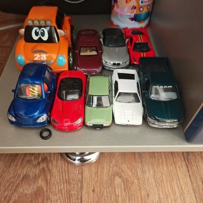 A shelf contains ten toy cars. The cars are arranged in a row, and they appear to be made of metal or plastic. The toy cars are of various colors and models, including an orange car with the number 23 printed on it, a maroon car, a silver car, a red sports car, a blue car, a red convertible car, a green car with Cyrillic letters on the bottom, a white car, and a dark green pickup truck with the word "RAM" on the windshield. A small black rubber ring lies on the shelf next to the blue car. The cars are located in the city of Pokrovskoye, Russia.