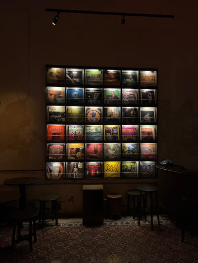 This image captures the interior of a dimly lit establishment, likely a bar, café, or listening room, located in Mérida, Mexico. The dominant feature is a large, square display on the left wall, showcasing 30 backlit vinyl record covers arranged in a 6x5 grid. Visible record titles include "SONORA SANTANERA," "ARETHA FRANKLIN SOUL'CO," "GO, MONGO! MONGO SANTAMARIA," "THE DAVE BRUBECK QUARTET," "QUINCY JONES THE WIDE WORLD OF," "MILES SMILES," and "It's Dance-Time WILLIE MITCHELL," indicating a diverse music selection with a strong emphasis on jazz, soul, and Latin genres.

To the right of the record display, several high-top tables with dark-legged stools are arranged, suggesting seating for patrons. The floor features a distinct, repeating geometric tile pattern in shades of red, brown, and white, characteristic of traditional architecture often found in Mérida. The walls have a textured, somewhat aged appearance, contributing to the venue's character. A single hanging light fixture with a warm glow is visible in the upper left, complementing the illumination from the record display. The overall atmosphere is intimate and cozy, indicative of an evening setting perfect for relaxing and enjoying music. No people are present, allowing the focus to remain on the meticulously curated decor and music collection.