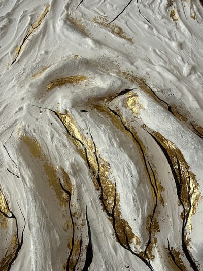 This is a close-up, abstract image of a textured surface with white, gold, and black elements. The white appears to be a thick, textured paint or plaster, with visible brushstrokes creating ridges and valleys. Interspersed within the white are irregular patches of shimmering gold leaf, applied in a somewhat fragmented manner. Thin, dark black lines, varying in thickness and intensity, are painted over the surface, often following the contours of the white texture and accentuating the gold leaf. Some areas show small flecks of gold scattered around. The lighting suggests a top-down source, casting shadows that emphasize the dimensionality of the surface. The image does not contain any recognizable objects, people, or specific environmental features, making it impossible to associate with a particular location in Užliekniai, Lithuania, or any other city.