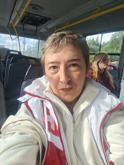 This is a portrait-oriented, close-up selfie taken from inside a bus. The primary subject is a person with short, light brown hair and brown eyes, who is wearing a white and red jacket. Their face is turned towards the camera, with a slight smile. Behind them, in the background, is another person with long blonde hair, wearing a dark red shirt. This person appears to be resting or looking away from the camera.

The setting is clearly a bus interior, with yellow poles and dark seats visible. The windows of the bus reveal a green, leafy landscape and a bright sky, suggesting it is daytime with fair weather. The sunlight is casting shadows and highlighting the subjects.

There is no visible text in the image. The overall mood appears casual and relaxed, possibly during a commute or a journey. The photo was taken in Gorka-Pokrovskaya, Russia.