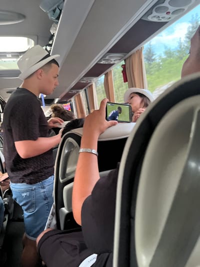 The image depicts the interior of a bus, likely while in motion, judging by the blurred background visible through the windows. The scene involves several people. A person in a white hat is visible in the foreground, looking upwards. Another individual is holding up a smartphone displaying a video, possibly of a soccer game. A third person, wearing a white hat, appears to be watching the video on the phone. The bus interior features overhead compartments, speakers, and window curtains. While the specific location within Verd, Slovenia, cannot be determined from the image alone, the presence of a bus suggests a public transportation context. The image captures a moment of leisure during travel, where individuals are engaged in entertainment on their devices.