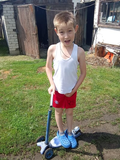 A young boy wearing a white tank top and red shorts stands on a blue three-wheeled scooter outdoors. He is looking at the camera with his mouth open. The background shows a rustic wooden shed with open doors, a brick wall, and a window on a white building. The ground is covered in grass and some patches of dirt. The scene appears to be in a rural or semi-rural setting, possibly in the Бородулиха ауылдық округі, Kazakhstan, given the context provided.