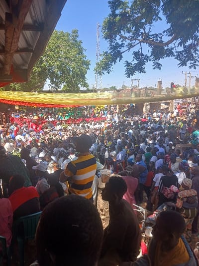 An outdoor scene in Gwolong, Nigeria, depicts a densely packed gathering of several hundred people under a clear blue sky. A large red and yellow fabric canopy stretches over a significant portion of the crowd, partially obscuring individuals seated or standing beneath it. In the immediate foreground, the back of an individual wearing a yellow and blue horizontally striped t-shirt and a dark cap is visible, arms crossed. To their left, another person in a red and black shirt sits on a green plastic chair. Partial views of additional attendees are visible in the foreground, including an individual in a dark brown shirt positioned lower right, looking towards the main gathering. A motorcycle is partially visible on the unpaved ground to the right. The background features mature trees with green foliage, a tall metallic telecommunications tower, and several incomplete or older concrete structures. The scene is brightly illuminated by natural sunlight, indicating daytime.