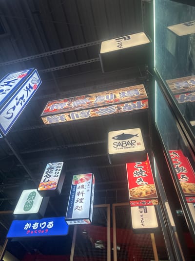 The image displays an indoor commercial area, identified as being in Clamart, France, characterized by numerous illuminated signs in Japanese script. These signs are suspended from a dark, industrial-style ceiling featuring exposed metal support rails. Prominently visible signs include one for "SABAR" with a black fish logo and the text "とろさば料理専門店" (specialized fatty mackerel cuisine restaurant). Other signs feature Japanese characters for establishments such as "かもりる" (Kamori-ru / Kyukyu), "こだわり の みたらし" (Kodawari no Mitarashi), and "磯料理処" (Iso Ryori-dokoro, seafood restaurant), some displaying images of food items like ramen, mackerel dishes, and griddled pancakes. The environment includes dark vertical walls, reflective panels on the right side showing reflections of signs and a vertical light strip, and a dark metal grid barrier in the lower background. Artificial lighting originates primarily from the illuminated signs. No individuals are present in the scene.