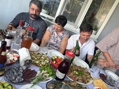 A medium shot captures a group of people sitting around a table laden with food and drinks, likely in a backyard or covered patio setting, judging by the open-air feel and the presence of a window with greenery visible beyond. The overall ambiance suggests a casual gathering or meal.
In the foreground, the table is a focal point, showcasing a variety of dishes. To the left, a bowl of salad with chopped tomatoes and white cheese is visible, next to a clear glass bottle containing a brown liquid. Next to that, a plate displays an assortment of sliced sausages and what appears to be cured meats. Further to the right, a platter holds roasted potatoes topped with a white cream or cheese. A bowl with sliced tomatoes and cucumbers sits centrally, adjacent to a large glass bottle with a red lid, containing a dark red beverage. In the lower right, another platter features cooked meat dishes garnished with fresh herbs, and slices of cheese are arranged on a yellow platter. The left side of the frame shows a dark-colored bottle with a yellow label and the neck of a lighter-colored bottle. Several plates and glasses are scattered across the table.
Four individuals are partially visible. On the left, a man with graying hair and a beard is seated, wearing a dark t-shirt. In the center, a woman with short dark hair is leaning forward, looking down. To her right, a man in a white shirt is holding a green bottle. On the far right, another person is partially visible, wearing a striped shirt. The hands of the people are also visible, with one man holding a bottle and another reaching towards the table.
The background reveals a window with a light-colored frame, and through it, potted plants are visible against a backdrop of foliage and dappled light, suggesting an outdoor or semi-outdoor environment. The walls appear to be a neutral gray color.
The scene is shot from a slightly elevated angle, looking down onto the table and the individuals. The lighting is natural, likely daylight, creating soft shadows. The overall impression is one of a relaxed, communal meal. The location mentioned, Яново-Грушевский, Russia, provides context for the cultural setting of the gathering.