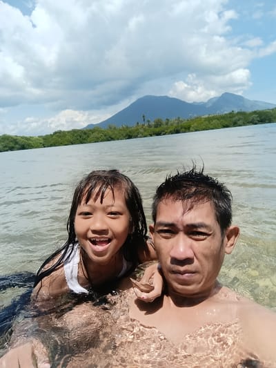 A man and a girl are captured in a selfie while waist-deep in the ocean. The girl, with wet hair, is smiling broadly. The man looks towards the camera with a more serious expression. In the background, the ocean stretches out to a line of trees and hills under a sky with scattered clouds. The water is clear enough to see beneath the surface near the shore.