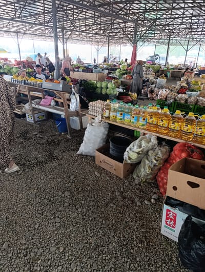 This is a medium shot, taken from ground level, of an outdoor market. The market is covered by a large, metal-framed roof, supported by pillars. The ground is covered with small grey rocks.
In the foreground and midground, there are several market stalls laden with produce and other goods. To the left, a woman wearing a leopard-print dress and white shoes walks away from the camera. In front of her is a stall with yellow fruit and baskets of vegetables. Further back, a man in a striped shirt is visible behind a stall of produce. To the right, there are stacks of vegetables, including onions and what appear to be cabbage. Several bottles of yellow cooking oil are lined up on a shelf. Boxes and sacks are scattered around the stalls, some containing produce and others appearing to be empty.
In the background, more market stalls and people are visible. Some individuals are interacting with vendors, while others are browsing the goods. The overall atmosphere appears to be busy and active. The market is open to the elements, with natural light filtering through the roof and the open sides. The environment suggests a bustling local marketplace. The city of Киров, Kyrgyzstan, is known for its agricultural markets, and this scene is consistent with the typical appearance of such a location.