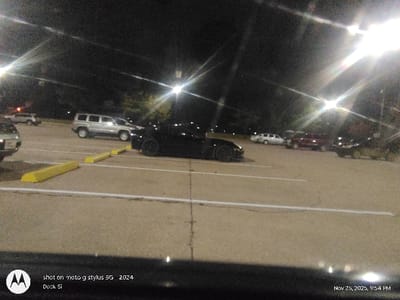 An aerial shot depicts an illuminated parking lot at night, dotted with several parked cars. The scene is captured with a timestamp of "Nov 25, 2025, 9:54 PM" and branded "shot on moto g stylus 5G 2024" and "Dock Si." Yellow parking barriers separate the lanes. Headlights illuminate the dark foreground, contrasting with the gray asphalt of the parking area.