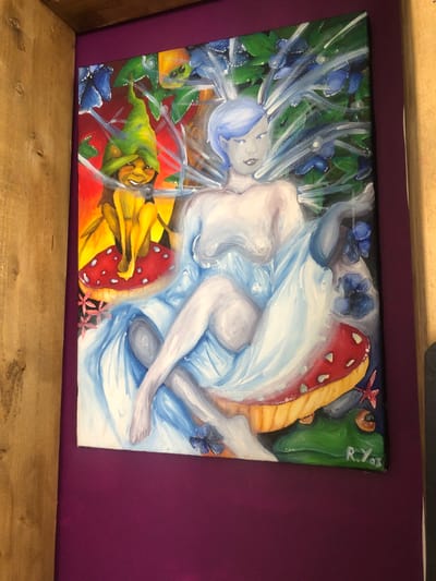 The image is a painting of a fairy with light blue hair sitting on a mushroom. She is draped in a flowing blue and white cloth. To her left, a small, grinning creature in a green pointy hat sits on another mushroom. The background is filled with green leaves and blue flowers, suggesting a woodland setting. The painting is signed "R.Y. '03" in the bottom right corner. The painting is mounted on a dark purple wall, with a wooden frame to the left. The overall impression is of a whimsical, magical scene. The provided image does not contain enough information to determine the location in Rother, United Kingdom.