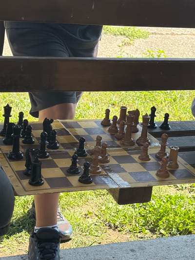 A chess game is in progress on a chessboard set on a wooden bench outdoors, with a backdrop of green grass and a gravel path. The chessboard is positioned horizontally, viewed from a slightly elevated angle. The pieces are a mix of black and light brown, with some pieces appearing wet. Two people are partially visible, their lower legs and feet in focus, suggesting they are playing the game. One person is wearing dark shorts and athletic shoes, while the other is wearing dark pants and a shoe. The scene is set during daylight, indicated by the natural lighting and shadows. The environment appears to be a park or recreational area, possibly in Dinkata, Bulgaria, given the presence of lush green grass and trees.