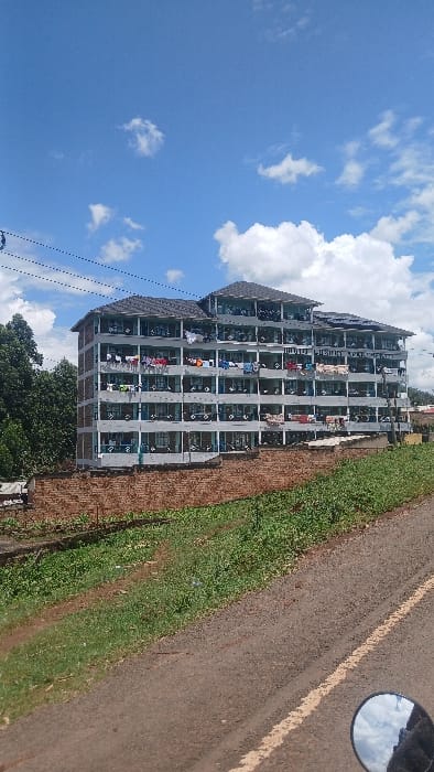 The image displays a multi-story residential or institutional building located in Boochi Tendere ward, Kenya, under a clear blue sky with scattered white cumulus clouds. The building, identified as "JUBILEE YOUTH CENTRE" by visible signage on its upper facade, is approximately five to six stories tall with a light grey painted exterior and a dark, multi-segmented tiled roof, on which solar panels are installed on the right side. Each floor features numerous windows, many secured with bars, and continuous balconies or verandas. A substantial amount of laundry, including various garments, towels, and blankets, is hanging to dry on railings and clotheslines across nearly all visible balconies.

The building is situated behind a red-brick retaining wall, with dense green trees and foliage partially obscuring its left side. In the immediate foreground, a dark dirt or gravel road extends horizontally, featuring a faded yellow line on its right side. A dirt path and green grass with low vegetation run alongside the road. A circular rearview mirror of a motorcycle, reflecting parts of the sky and building, is present in the lower right corner of the frame, indicating the photographer's perspective. Black power lines with multiple strands traverse the upper left portion of the image. No individuals are depicted within the scene. The environment indicates bright daylight conditions.