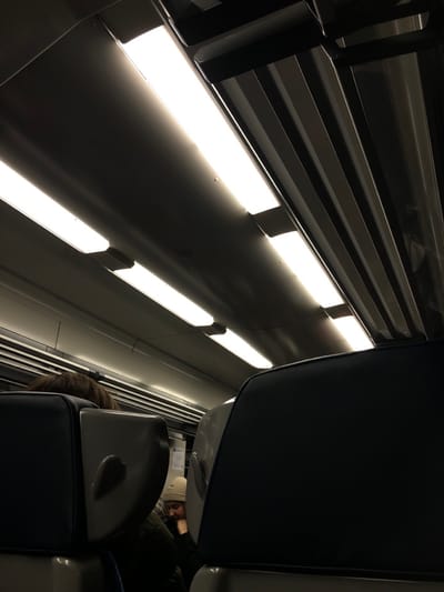 The interior of a train car shows overhead lights and luggage racks. Seats with blue upholstery face forward. A passenger in the distance wears a white hat and is reading. The lighting is bright, casting harsh shadows on the walls.