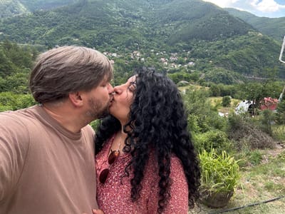 This media file is a close-up, rotated, outdoor photograph of a couple kissing. The subjects are a man and a woman, positioned in the foreground with their faces touching. The woman has long, dark, curly hair and is wearing a red floral dress. A pair of sunglasses rests on her shoulder. The man has greying hair and is wearing a brown t-shirt. Their focus is on each other, with an expression of affection.

Behind the couple, a lush, green, mountainous landscape unfolds. The scene is dotted with houses and trees, suggesting a rural or semi-rural setting. The sky above is partly cloudy. The overall atmosphere conveyed is one of intimacy and romance set against a beautiful natural backdrop. The time of day appears to be daytime, with bright natural light. The location is identified as Tompsan, Bulgaria. There is no visible text in the image.