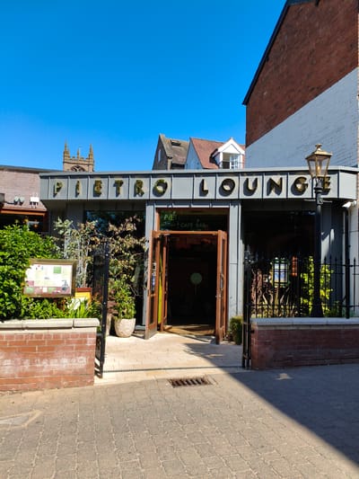 The image shows the exterior of "PIETRO LOUNGE CAFE BAR" located in the city of Ludlow, United Kingdom. The building features a modern facade with large, golden lettering spelling out "PIETRO LOUNGE". The entrance has double wooden doors, which are open, revealing a glimpse of the interior. Several potted plants and greenery are strategically placed outside the cafe, along with a brick planter box and a framed menu displayed on an easel. A black wrought-iron fence surrounds a portion of the cafe's exterior. In the background, a church spire is visible against a clear blue sky, suggesting the cafe is situated in an area with historical architecture. The ground in front of the cafe is paved with cobbles and brickwork. A vintage-style lamppost stands to the right of the entrance. The lighting indicates it is daytime, with sunlight casting shadows on the ground.