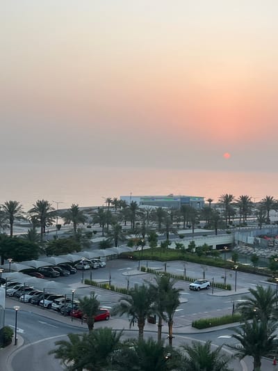 This aerial, wide-angle shot captures a serene sunset over a coastal city, likely Ras Al-Khaimah, United Arab Emirates. The sun, a soft orange-red disc, hangs low in a hazy, peachy sky, casting a warm glow over the scene. The landscape is a blend of natural and urban elements.
To the left, the ocean stretches out to the horizon, its surface a muted grey under the diffused light. The coast is lined with lush palm trees, their fronds creating a rhythmic pattern against the sky. Beyond the trees, modern architecture is visible, including a multi-story building with a blue and white facade, featuring prominent signage that reads "Ras Al-Khaimah" in English and Arabic, along with a green logo.
To the right, a well-maintained parking area is visible, filled with a variety of cars, mostly white and grey, parked under white canopies. The road curves gently, with streetlights casting pools of light. The presence of palm trees, modern buildings, and a coastal setting are strong indicators of the United Arab Emirates. The overall mood is tranquil and picturesque, characteristic of a calm evening by the sea. The image appears to be taken from a high vantage point, possibly a hotel room or a tall building overlooking the city and coastline. The weather appears to be clear with a touch of atmospheric haze, typical for the region during sunset.