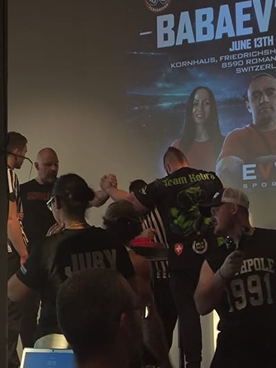 The image depicts an indoor arm wrestling event, likely at the Kornhaus in Romanshorn, Switzerland, as indicated by the text on the screen. In the foreground, several individuals are visible, some actively participating or spectating. One person, wearing a black shirt with "JURY" printed in large grey letters on the back, is positioned in the lower center of the frame. To their left, a person in a black t-shirt with "GORILL" printed on it, is engaged in arm wrestling with another individual whose arm is not fully visible. Further to the right, a man in a cap and headphones is looking to the side. In the background, a large screen displays promotional material for an event featuring the name "-BABAEV" and event details including "JUNE 13TH KORNHAUS, FRIEDRICHSHOF, 8590 ROMANSHORN, SWITZERLAND." The screen also shows images of two individuals, a woman and a man. The lighting suggests an indoor venue with artificial lighting.