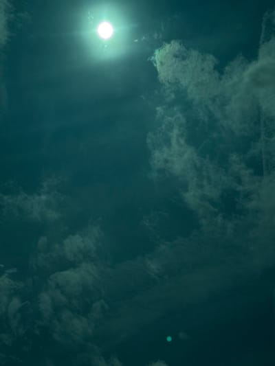This image depicts an atmospheric scene over Longeville-lès-Saint-Avold, France. The upper central area features a brilliant, circular white light source, characteristic of the sun, exhibiting prominent lens flare artifacts, including radiating light beams. The sky presents a deep teal coloration. Varied formations of diffuse, light-colored clouds are distributed throughout the frame, with heavier concentrations observed along the right vertical edge and in the lower left quadrant. Two distinct, circular, blue-green light anomalies are present in the lower center. A subtle, vertical linear artifact extends along the far right edge of the image. The scene contains no visible human subjects, structures, or terrestrial features, indicating an unobstructed aerial or elevated perspective.