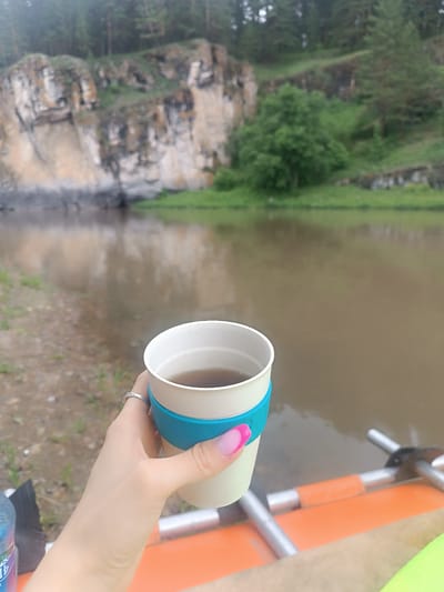 In Idrisovo, Russia, a woman's hand with a silver ring and pink-tipped fingernails holds a light-colored reusable cup with a turquoise silicone sleeve, filled with a dark liquid. The cup is positioned in the foreground, obscuring part of an orange inflatable object with silver metal components, possibly part of a boat or raft. Behind the hand and cup, a wide, muddy river flows, reflecting the overcast sky. The riverbank to the left is composed of brown soil and sparse green vegetation. In the background, a steep, rocky cliff face rises, partially covered with green moss and lichen. Above the cliff, a dense forest of pine trees stretches across the landscape under a hazy sky.
