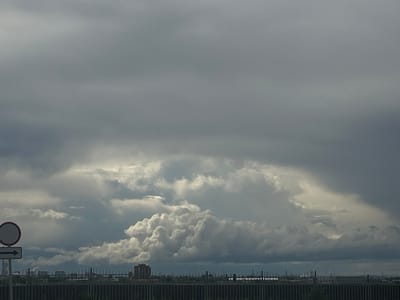 The image shows an overcast sky with a large formation of cumulonimbus clouds. In the foreground, there is a road sign with a white circle and a horizontal arrow pointing to the right. Behind the sign, a fence runs across the lower part of the frame, obscuring the horizon. Beyond the fence, there is a cityscape with buildings and industrial structures. Given the location in Тимофеевка, Russia, the industrial features suggest a possible urban setting. The absence of people and the focus on the environmental and man-made elements create a stark, descriptive portrayal of the scene.