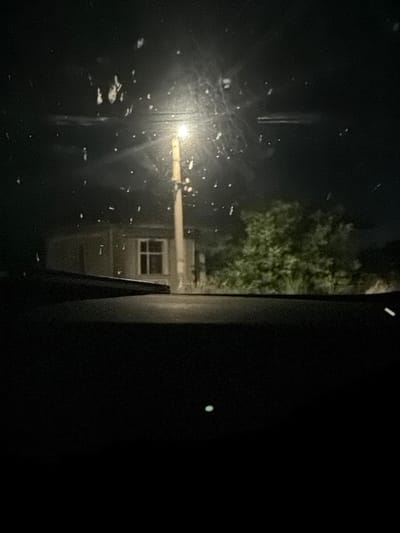 The image captures a nighttime scene from inside a vehicle looking through the windshield, which is speckled with raindrops or water spots. A street light illuminates a utility pole situated in front of a building with a visible window. To the right of the building and utility pole, dense foliage is present. The foreground is dominated by the dark interior of the vehicle, with a hint of the dashboard visible. The overall environment suggests an outdoor setting in a location that appears to be Smyadovo, Bulgaria, given the context. No individuals or specific actions are discernible, but the scene implies a stationary or slow-moving perspective, possibly from a parked car or a vehicle traveling at low speed.