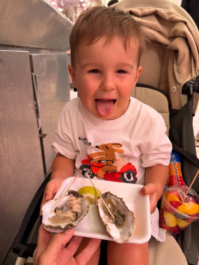 A young child with an open mouth and tongue sticking out is seated in a stroller. The child wears a white t-shirt with a graphic print. In front of the child, held by an adult's hands, is a white tray with two oysters, one of which has a toothpick inserted. A slice of lemon is also on the tray. To the right of the tray is a clear plastic cup filled with cut fruit, including strawberries, peaches, and blueberries, also with a toothpick inserted. The background suggests an indoor setting with metallic surfaces and a light-colored fabric, possibly a market or food stall. The child is in the city of Alboraia/Alboraya, Spain.