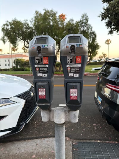 Two parking meters are shown in Laguna Beach, California, USA. They are silver and gray, with displays at the top showing "00:03". Below the display are slots for coins and credit card readers. One meter has a sticker that says "THIS METER SERVES THE SPACE ON YOUR LEFT" and the other says "THIS METER SERVES THE SPACE ON YOUR RIGHT". Both meters have a sticker indicating "METERS ENFORCED EVERYDAY 8:00 A.M. - 7:00 P.M." and a "10 HOUR PARKING LIMIT until 7:00 P.M." A white car is parked to the left of the meters, and a dark SUV is parked to the right. The background shows trees and a building, with a clear sky indicating it is daytime. The meters are mounted on a gray pole.
