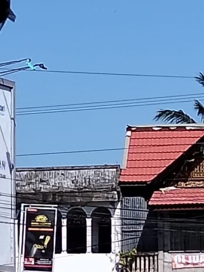 The scene shows a street view with buildings and power lines under a bright blue sky. A two-story structure with a weathered gray exterior and three arched windows sits beside a building with a red-tiled roof and detailed facade. A sign with a black and yellow design is visible in front of the gray building. A few palm fronds are visible behind the building with the red roof. Power lines stretch horizontally across the upper part of the frame.