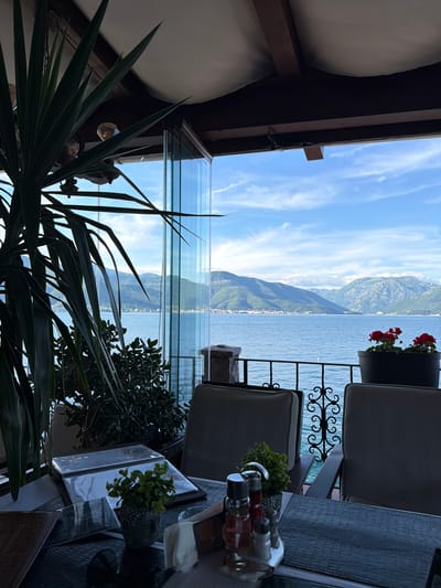 The image depicts a partially obscured view from a restaurant or balcony in Krašići, Montenegro, facing the Bay of Kotor. A mountain range dominates the distant backdrop. The foreground features interior elements including a glass partition, seating, plants, and table settings with condiments. Sunlight illuminates parts of the scene.