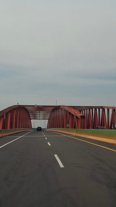 The image depicts a highway leading towards a large, red truss bridge under an overcast sky. The perspective is from a vehicle traveling on the road, which is marked with white lane lines. Two cars are visible on the bridge, one in the left lane and another further ahead in the right lane. The bridge's red metal structure is prominent, with its complex lattice of beams and supports. On the right side of the bridge, a sign indicating a speed limit of 40 is visible. The surroundings beyond the bridge are not clearly discernible due to the distance and atmospheric conditions. The image captures a moment of transit on a modern highway, with the imposing bridge as the central architectural element. The scene is devoid of any discernible landmarks that would specifically place it within the city of Stabat, Indonesia; the image primarily focuses on the bridge and the roadway.