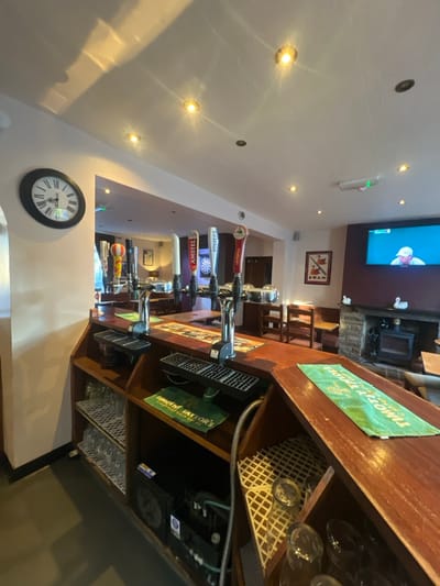 A bar area featuring multiple beer taps labeled with brands like "Luintenica," "Fosters," and "Amstel." Above the taps, a television displays content, and a framed picture hangs on the wall. Behind the bar counter, Timothy Taylor's signage is visible. Glasses are stored neatly on shelves, and a clock is positioned on the wall below the taps. The room is brightly lit with recessed lighting.