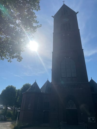 The image captures a view of a brick church tower and adjacent building, likely a church, under a bright, sunny sky. The sun is intensely bright, causing lens flare that streaks across the image, suggesting it is midday. The sky is a clear blue with some wispy white clouds. Lush green trees are visible in the foreground and to the right of the church, with one tree prominently positioned on the left, its leaves illuminated by the strong sunlight.
The church tower is tall and made of dark brick. It has several arched windows and what appears to be a clock face or a circular emblem with a blue background and a figure in the center, near the top. A cross is visible at the very apex of the tower. The main body of the church, also brick, features arched windows and ornate brickwork detailing. A smaller, gabled section of the church is visible to the right.
The scene is set outdoors, likely in a town or village, given the presence of the church and trees lining a path or road. A white sign with some text is visible near the base of a lamp post on the right side of the frame. The overall mood is bright and serene, typical of a sunny day. The orientation of the image is rotated 90 degrees counter-clockwise. No people are visible in the frame.