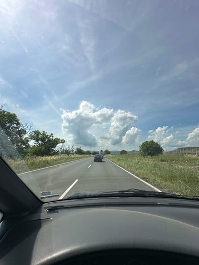 The image captures a view from the interior of a car driving on a road in Sadievo, Bulgaria, on a cloudy day. The road ahead is paved and marked with white lines, with a few cars visible in the distance. The roadside is lined with green grass and trees, with rolling hills and a fence in the background. The sky is a mix of blue and white, with large, fluffy clouds creating a dramatic effect. The car's dashboard and windshield are visible in the foreground, indicating the perspective of the person taking the photo. The overall scene suggests a journey through a rural landscape under a dynamic sky.