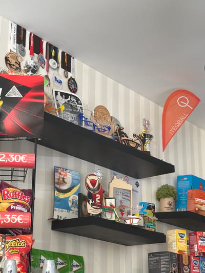 The image depicts an indoor commercial or club setting, likely a cafe or shop associated with the sport of Teqball, located in the Santarém region of Portugal.
The upper section of the scene features a white ceiling and walls covered with light-colored, vertically striped wallpaper. Three black floating shelves span horizontally across the wall.
On the top shelf, numerous medals, some labeled "OPEN TEQBALL" and "FREGUESIA," hang from black felt strips. Various acrylic and wooden trophies and plaques are displayed, many featuring stylized figures of Teqball players. Text on these awards includes "TEQBALL," "Federação Teqball Portugal," and mentions specific Portuguese locations such as "Santarém," "Abrantes," "Almeirim," "Sintra," and "Cad Coruche." A small, orange teardrop-shaped flag with the "TEQBALL" logo stands on the right side of this shelf. A red and black poster with a QR code is partially visible on the far left.
The middle shelf showcases additional Teqball awards, including plaques for "SINTRA" and "Dobles Mix," and a "2º Doubles" award. A small potted artificial plant labeled "madremaria sweet home" sits on the right, next to retail boxes of Delta Decaf coffee and Cappuccino Classic.
The bottom portion of the image displays retail products on a black wire rack. Bags of Ruffles (Sabor a Presunto) and Cheetos (Sticks Palitos) are visible with red price tags showing "2,30€" and "1,35€." Below these, rolls of Trident gum are neatly stacked. On the far right, additional retail boxes of Tetley tea (Immune, Boost, Fruity Infusion) and various coffee products are arranged.
No people are visible in the scene. The items displayed suggest an establishment with a strong connection to Teqball events and achievements within the Santarém district and surrounding areas of Portugal, possibly functioning as a local sports club, academy, or a cafe/shop that celebrates and supports the sport.