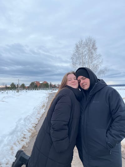 The image portrays two individuals, a male and a female, posing closely together outdoors in Kalinino, Russia, during winter conditions. The female, on the left, has long brown hair and wears a black quilted puffer coat and dark, thick-soled boots. She looks towards the camera with a subtle smile, her head resting against the male's. The male, positioned on the right, wears a black hooded jacket featuring an "OUTVENTURE" logo on the left sleeve, and a black beanie, with his head leaning against the female's.
The ground is blanketed in snow, with a cleared, dark-colored path running diagonally from the lower-left to the center-right, bordered by snowdrifts. In the background on the left, residential buildings with orange-red roofs are visible amidst bare trees and fences, alongside utility poles and power lines. To the right, a prominent bare-branched tree with light bark stands against a backdrop of a vast, snow-covered field extending to the horizon. The sky is uniformly overcast with multiple layers of grey clouds.
