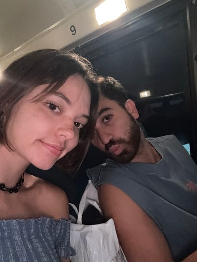 Two individuals, a woman and a man, are pictured in close proximity, likely on a form of public transportation given the overhead lighting and a visible number "9" on a wall. The woman, positioned in the foreground on the left, has short, dark hair and is looking towards the camera with a slight smile. She is wearing an off-the-shoulder blue top and a dark beaded necklace. The man, to her right, is looking towards the camera as well, with dark hair and a beard. He is wearing a grey t-shirt. In the background, there are dark surfaces and what appears to be a white bag. The overall environment suggests an indoor setting, possibly a train or bus, during nighttime or low-light conditions. The location is identified as Halfway, United States of America.