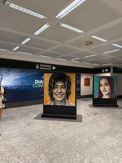The image depicts an indoor public space, likely a transportation hub such as an airport or train station, given the "DEPARTURES" sign in Italian and English. Two large, vibrant mosaic portraits are displayed prominently on stands. The portrait on the left is of a person with dark, curly hair and a wide smile. The portrait on the right is of a young woman with long, dark hair and a neutral expression. Both portraits are composed of small, colorful pieces that create a pixelated effect.
In the background, a large advertisement featuring a seascape with crashing waves and a stylized depiction of a water bottle or glass is visible. Another smaller artwork, a framed painting with an orange hue, is also present on the wall. The ceiling features a grid of fluorescent lights and ventilation grilles. The floor is made of speckled granite tiles. A woman with a backpack and a white dress is partially visible on the left side of the frame, appearing to walk away from the viewer. The overall environment suggests an exhibition or art installation within a transit area.