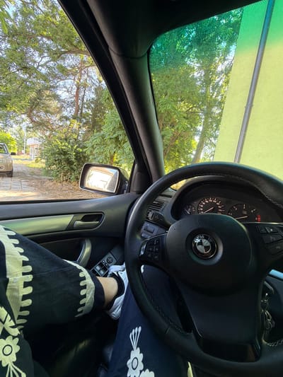 The image shows the interior of a car, with a focus on the steering wheel and dashboard. The driver's leg is visible, wearing black pants with a white floral pattern and white sneakers. The steering wheel is black leather with stitching and has a BMW logo in the center. The dashboard displays a speedometer and tachometer, with the car's interior appearing to be in good condition.

Outside the car, through the windows, lush green trees and foliage are visible, suggesting a location surrounded by nature. The sky is partly cloudy and bright, indicating daytime. A light brown car is parked on a paved driveway in the distance. The car's side mirror reflects a building or house. The overall scene suggests a serene drive through a scenic area. The image is taken from the driver's perspective, implying the person is either driving or about to drive. The location is identified as the city of Dinkata, Bulgaria.