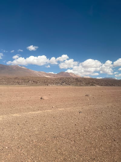 A panoramic shot reveals a vast, dry landscape. The ground, covered in small rocks, transitions into a mountain range where a cloud-filled sky is visible above. The sky is a bright blue. The clouds are white and puffy.