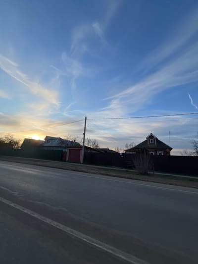 The image presents a roadside view under a partly cloudy sky. A road runs along the right side of the frame. Adjacent to the road, a row of buildings, including a distinctive house with a gable roof and another building with a red-painted area, are visible. Utility poles and wires stretch across the sky above the buildings. The lighting suggests either sunrise or sunset, with the sun low on the horizon.

The location is not explicitly visible from the photo itself; Yasenevo is mentioned in the prompt but not visually present.