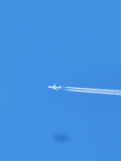 An airplane is flying through a bright blue sky, leaving a white contrail behind it. The plane is small and white, appearing as a silhouette against the sky. The contrail is long and wispy, stretching out horizontally across the frame. The image appears to be taken from a ground-level perspective, looking up at the plane. There is a faint, dark shadow in the lower portion of the image, likely cast by an object or obstruction not visible in the frame. The image does not contain any information about the city of Zgorigrad, Bulgaria.