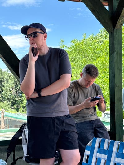 This media file depicts two men relaxing outdoors on a sunny day, likely under a wooden pergola or structure. The image is rotated 90 degrees counter-clockwise.
In the foreground, a man with a dark baseball cap, sunglasses, and a light beard is holding a black smartphone to his left ear, appearing to be on a call. He is wearing a dark grey t-shirt and black shorts, with a black smartwatch visible on his left wrist. A black backpack is visible near his legs on the ground.
Behind him and to his right, another man with short dark hair, wearing a light olive-colored t-shirt and dark trousers or long shorts, is looking down at a smartphone held in both hands.
The background features a bright blue sky with scattered white clouds on the left side. Lush green trees and foliage fill most of the upper and right background, suggesting a natural or park-like setting. Parts of a wooden beam structure are visible in the upper and left portions of the frame. On the far right, a blue and white striped chair is partially visible. The overall impression is one of leisure and relaxation in an outdoor environment, during daylight hours, in the Grocka Urban Municipality, Serbia. There is no visible text in the image.